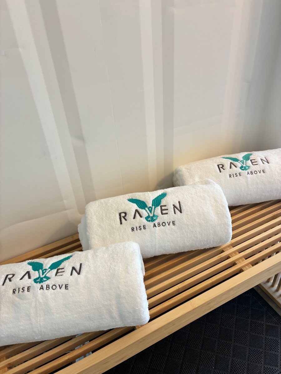 Rolled guest towels embroidered with the Raven logo on a wooden bench